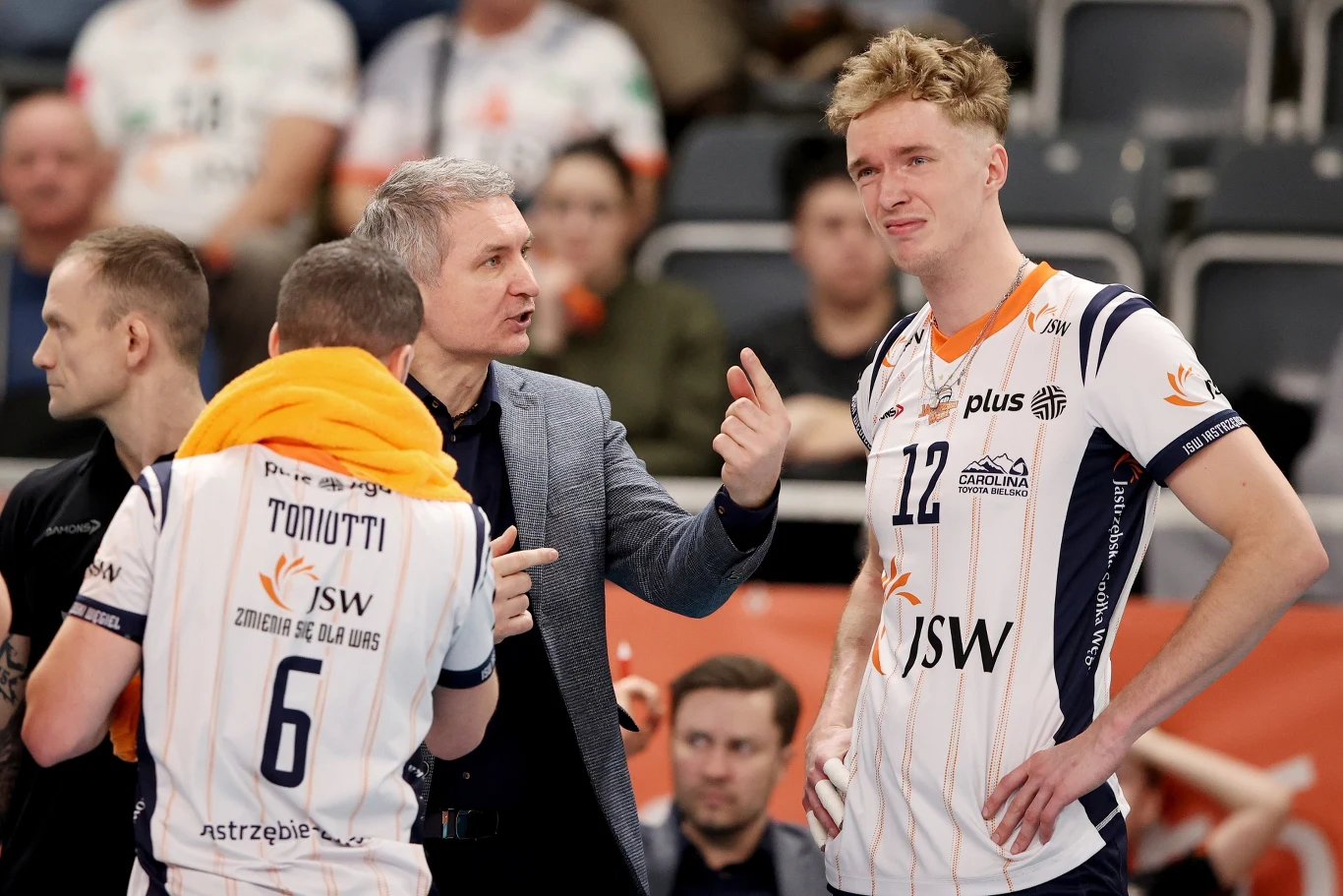 Trener drużyny siatkarskiej rozmawia z zawodnikami w trakcie przerwy podczas meczu, zawodnicy ubrani w klubowe stroje z widocznymi nazwiskami i numerami, w tle publiczność na trybunach.