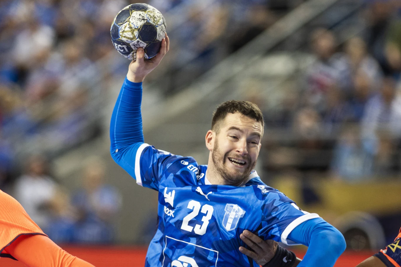 Zawodnik drużyny piłki ręcznej w niebieskim stroju z numerem 23 na koszulce unosi piłkę do rzutu podczas meczu, wokół niego widoczni są inni gracze oraz rozmyta publiczność na trybunach.