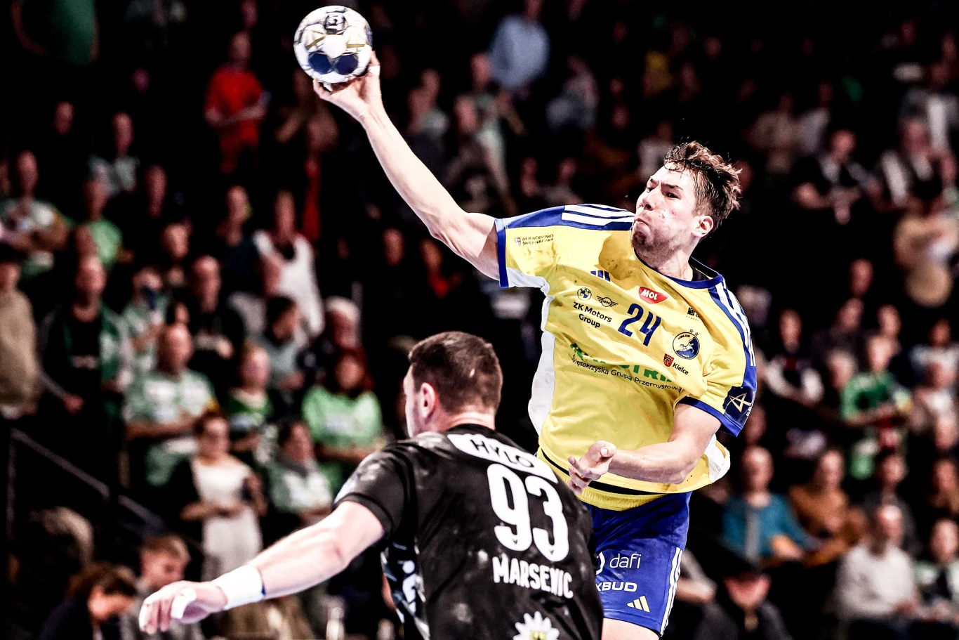 Zawodnik w żółto-niebieskiej koszulce wykonuje dynamiczny rzut piłką ręczną, unosząc się nad przeciwnikiem w czarnej koszulce na arenie sportowej, kibice w tle obserwują akcję.