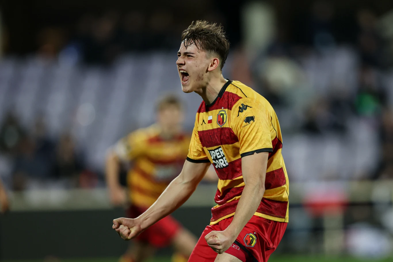 Młody mężczyzna w żółto-czerwonym stroju sportowym wyraża intensywne emocje po zdobyciu bramki na stadionie, w tle rozmyci kibice oraz inny zawodnik.