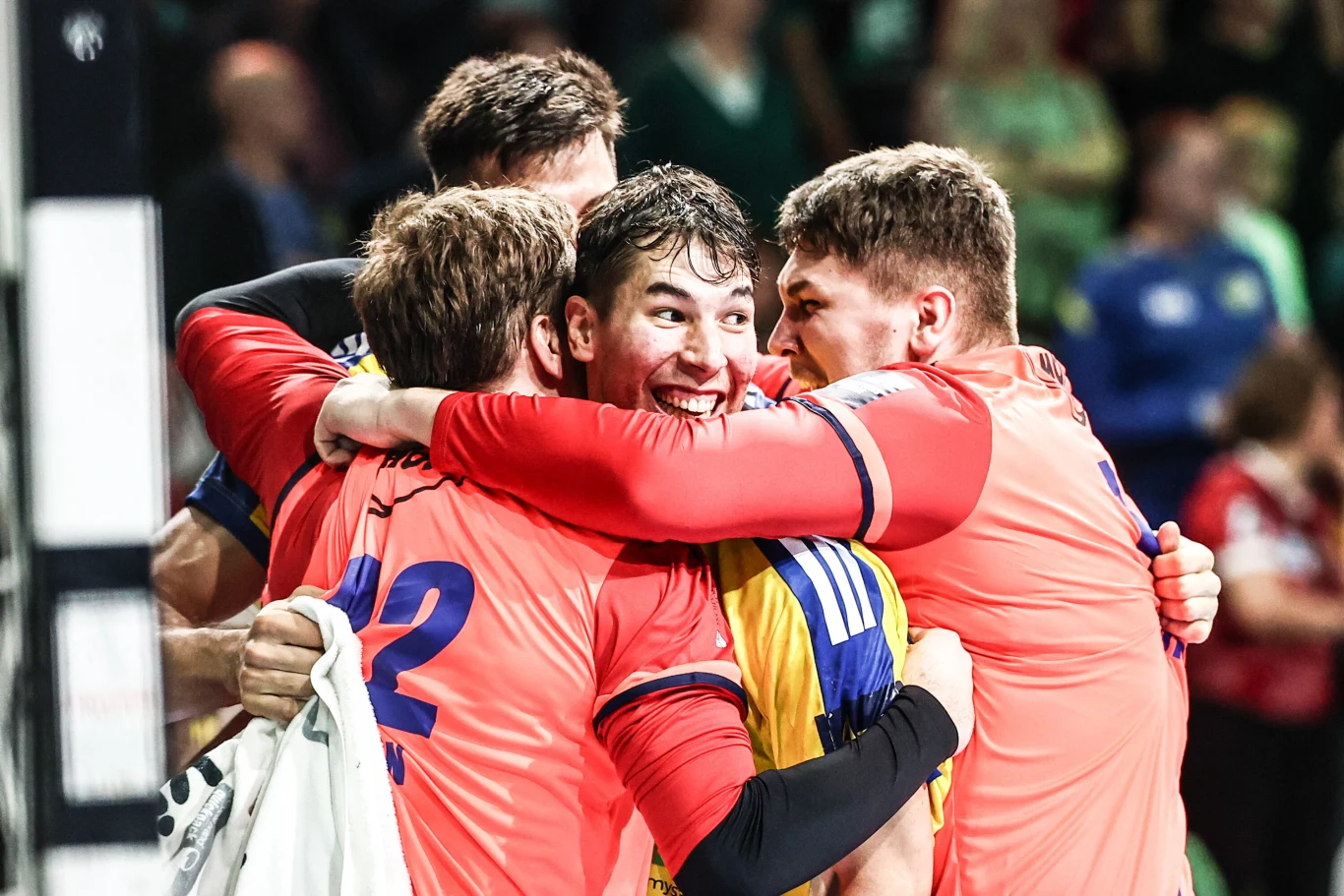 Grupa sportowców w kolorowych strojach ściska się z radości na tle rozmytego tłumu, jeden z zawodników uśmiecha się szeroko, wyrażając silne emocje po wygranym meczu.