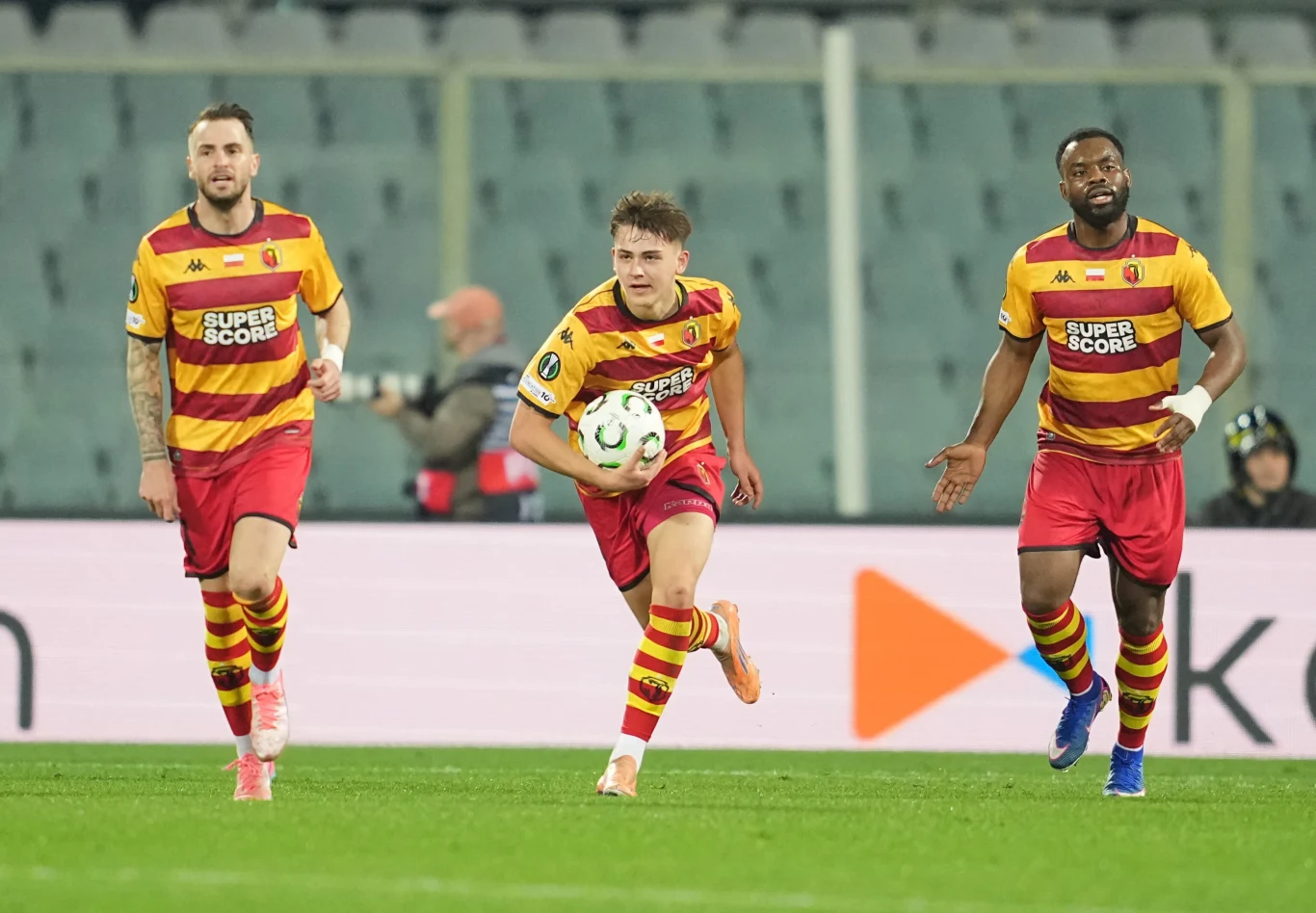 Trzech piłkarzy w żółto-czerwonych strojach z piłką podczas meczu na stadionie, w tle trybuny z widownią.