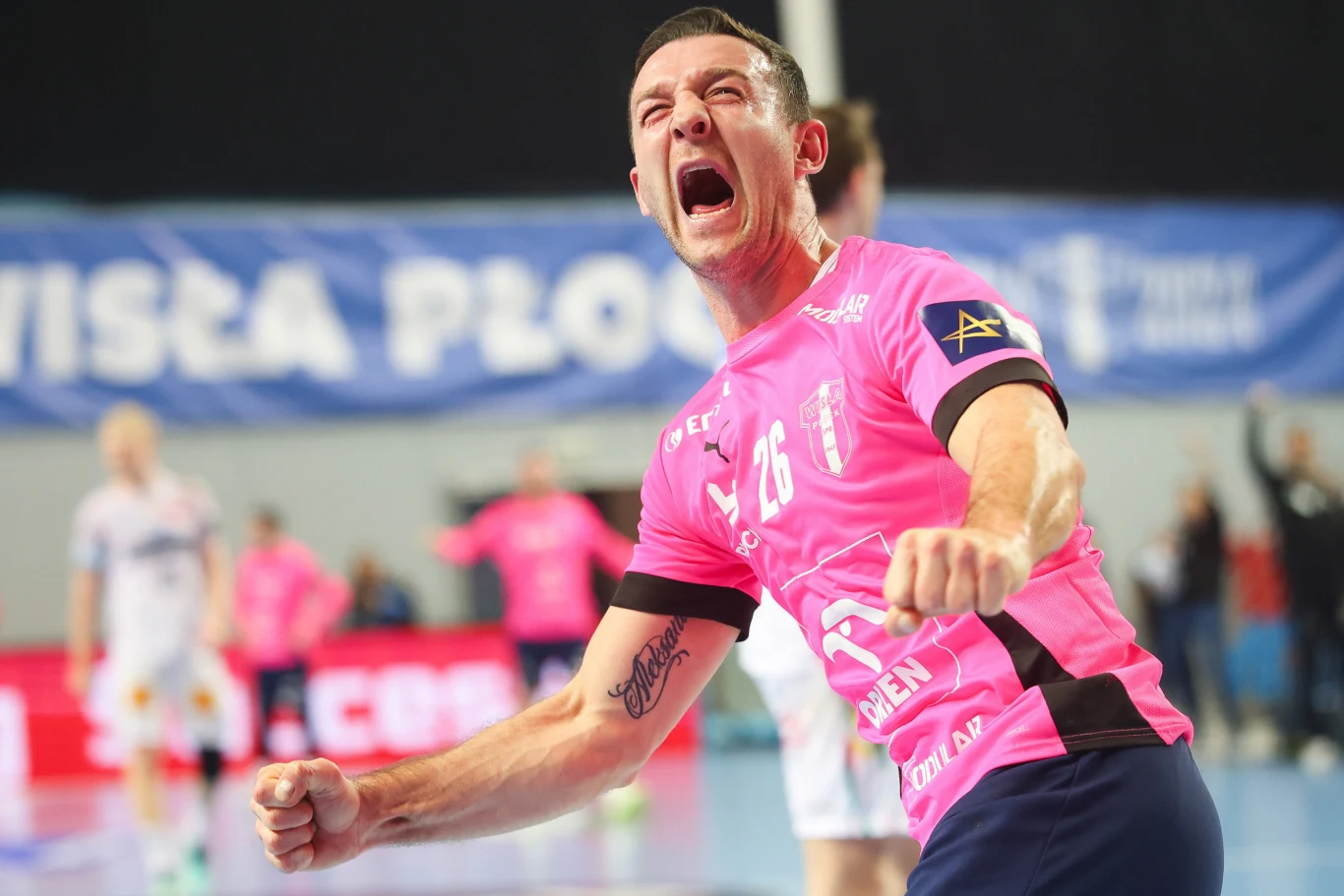 Zawodnik drużyny piłki ręcznej ubrany w różowy strój celebrowający sukces, z zaciśniętymi pięściami i wyrazem radości oraz triumfu na twarzy, w tle inni zawodnicy i nieostre napisy na banerach.