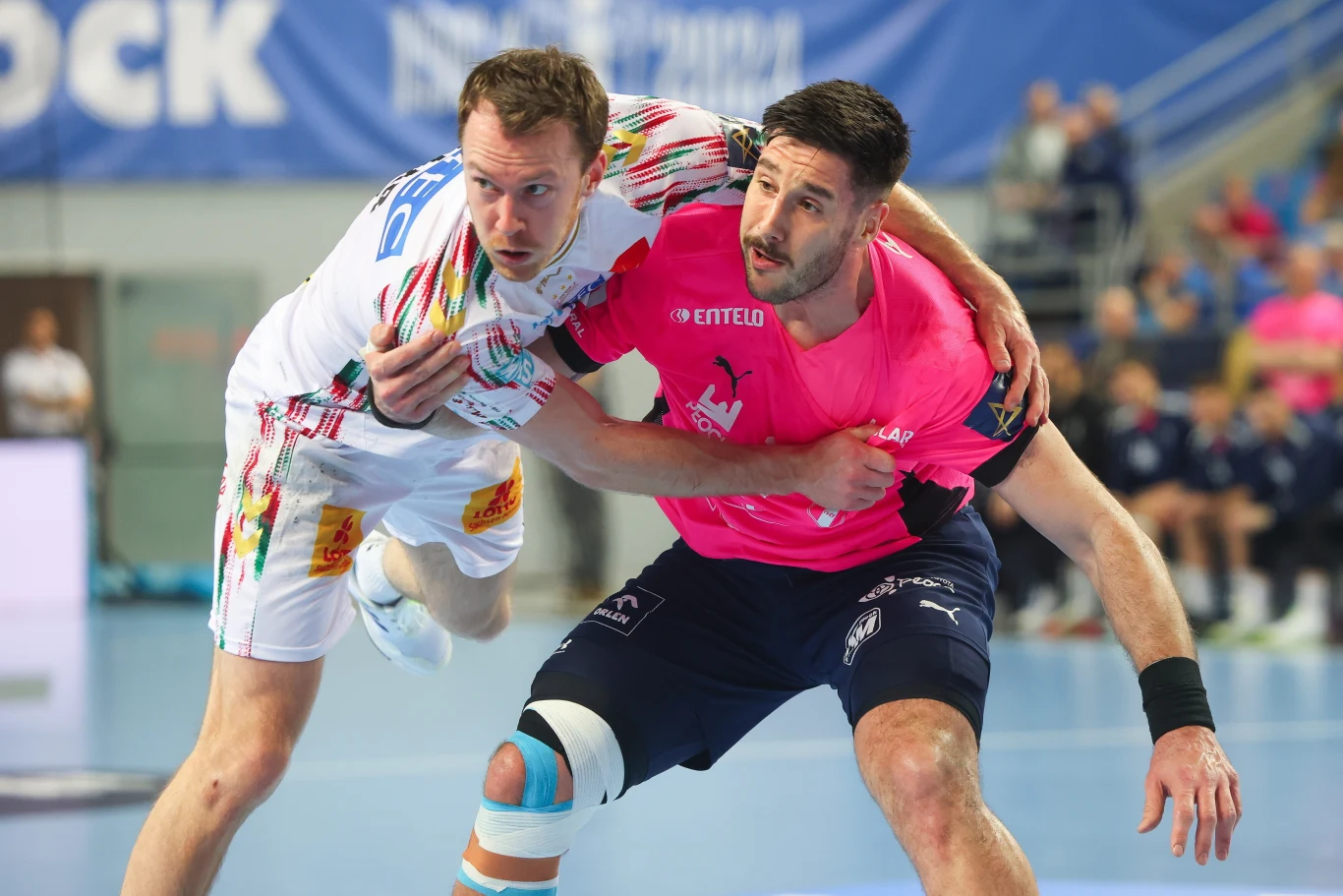 Dwóch szczypiornistów walczy o piłkę podczas intensywnego meczu halowego, ubrani w kontrastowe stroje drużynowe, na tle rozmazanej publiczności i niebieskiej podłogi sportowej.