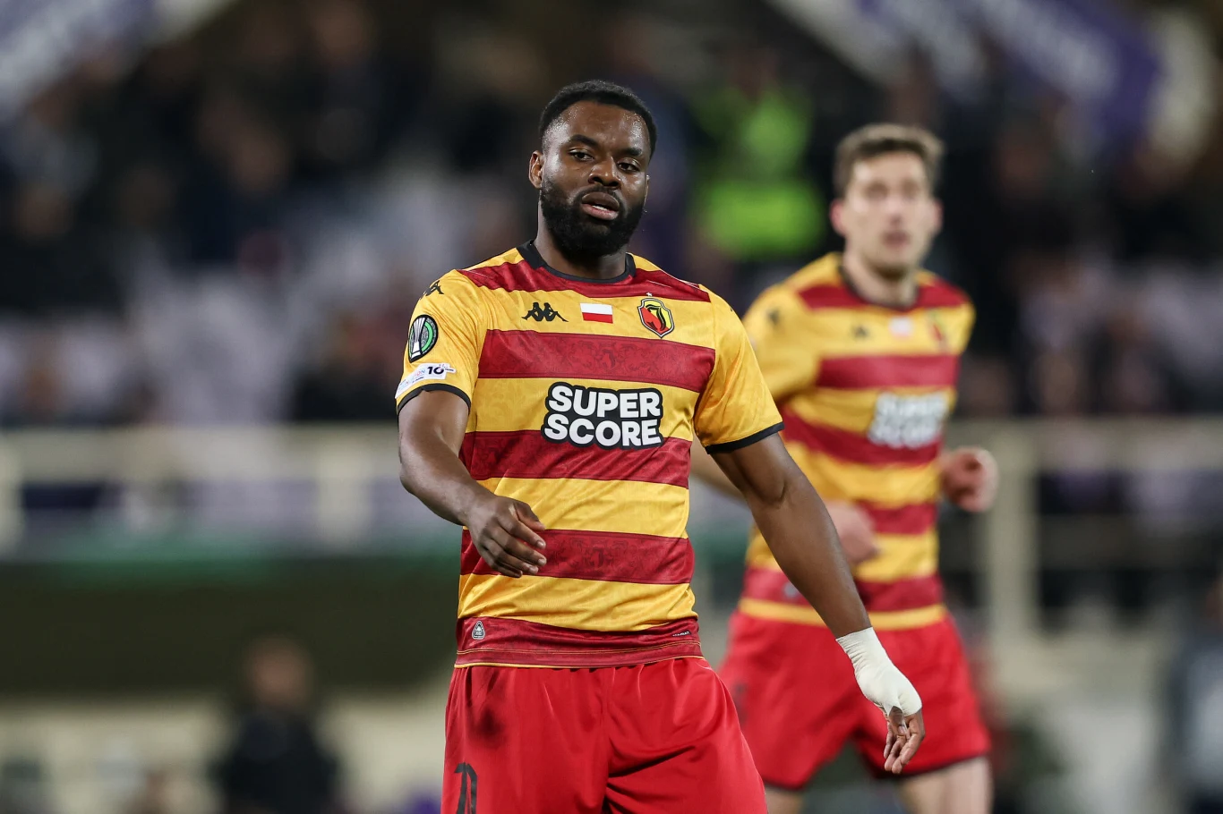 Piłkarz w pasiastym żółto-czerwonym stroju drużyny z napisem 'Super Score' na koszulce stoi na boisku podczas meczu, w tle widoczny inny zawodnik.