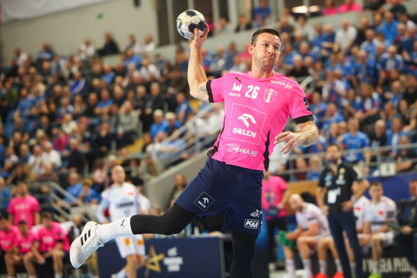Zawodnik piłki ręcznej w różowej koszulce z numerem 26 w dynamicznym ruchu podczas rzutu, w tle widać publiczność oraz innych zawodników i trenerów ubranych w stroje sportowe.