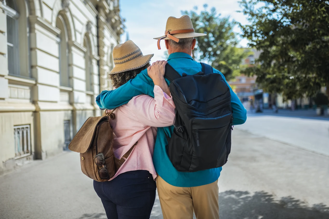 Zamieniają mieszkania na życie w podróży Para starszych osób spaceruje razem po mieście, trzymając się za plecy; oboje mają kapelusze, plecaki oraz wygodne ubrania turystyczne.
