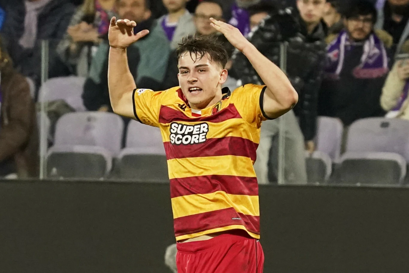 Młody piłkarz w żółto-czerwonej koszulce z napisem Super Score wyraża emocje podczas meczu piłkarskiego, w tle widoczni kibice na trybunach.