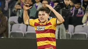 Młody piłkarz w żółto-czerwonej koszulce z napisem Super Score wyraża emocje podczas meczu piłkarskiego, w tle widoczni kibice na trybunach.