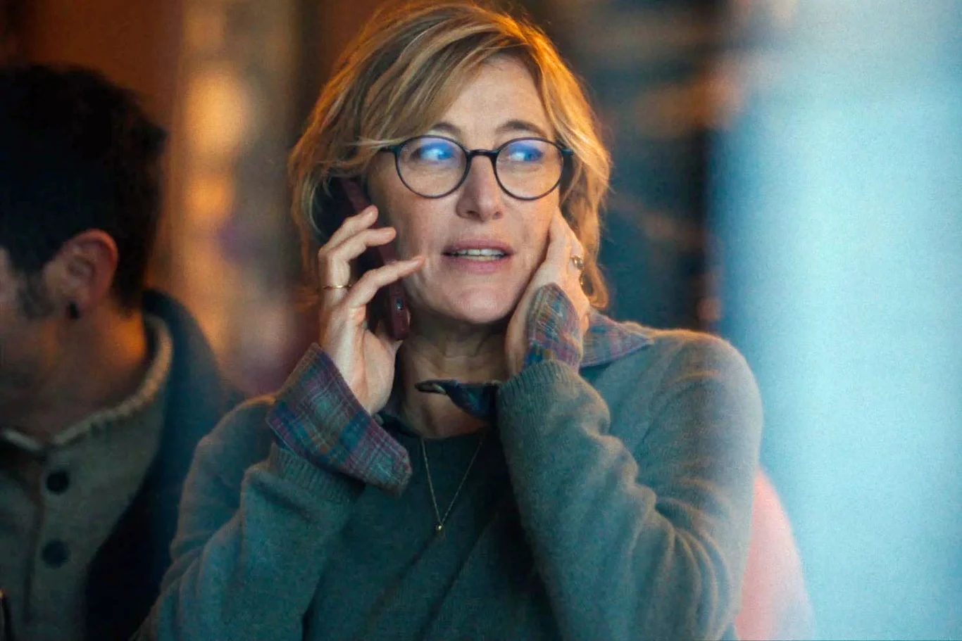 Valeria Bruni Tedeschi w scenie z filmu "L'Attachement" Kobieta w okularach rozmawiająca przez telefon, ubrana w luźny sweter, z wyrazem zamyślenia na twarzy, znajdująca się w jasnym pomieszczeniu, w tle częściowo widoczna inna osoba.