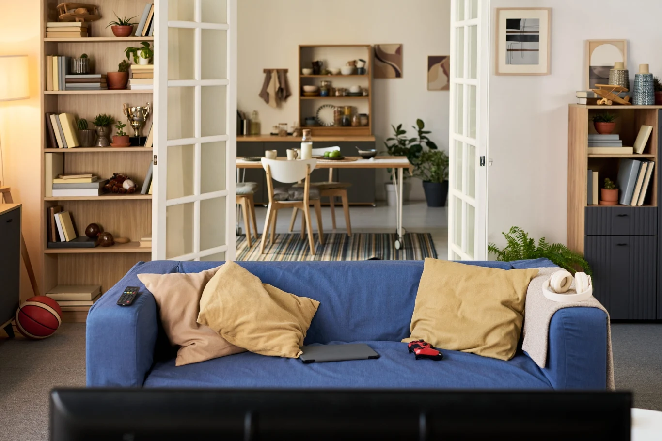 Salon z niebieską sofą, na której leżą poduszki, laptop i kontroler do gier, w tle widoczna otwarta przestrzeń prowadząca do jadalni z jasnymi meblami i półkami pełnymi książek oraz roślin.