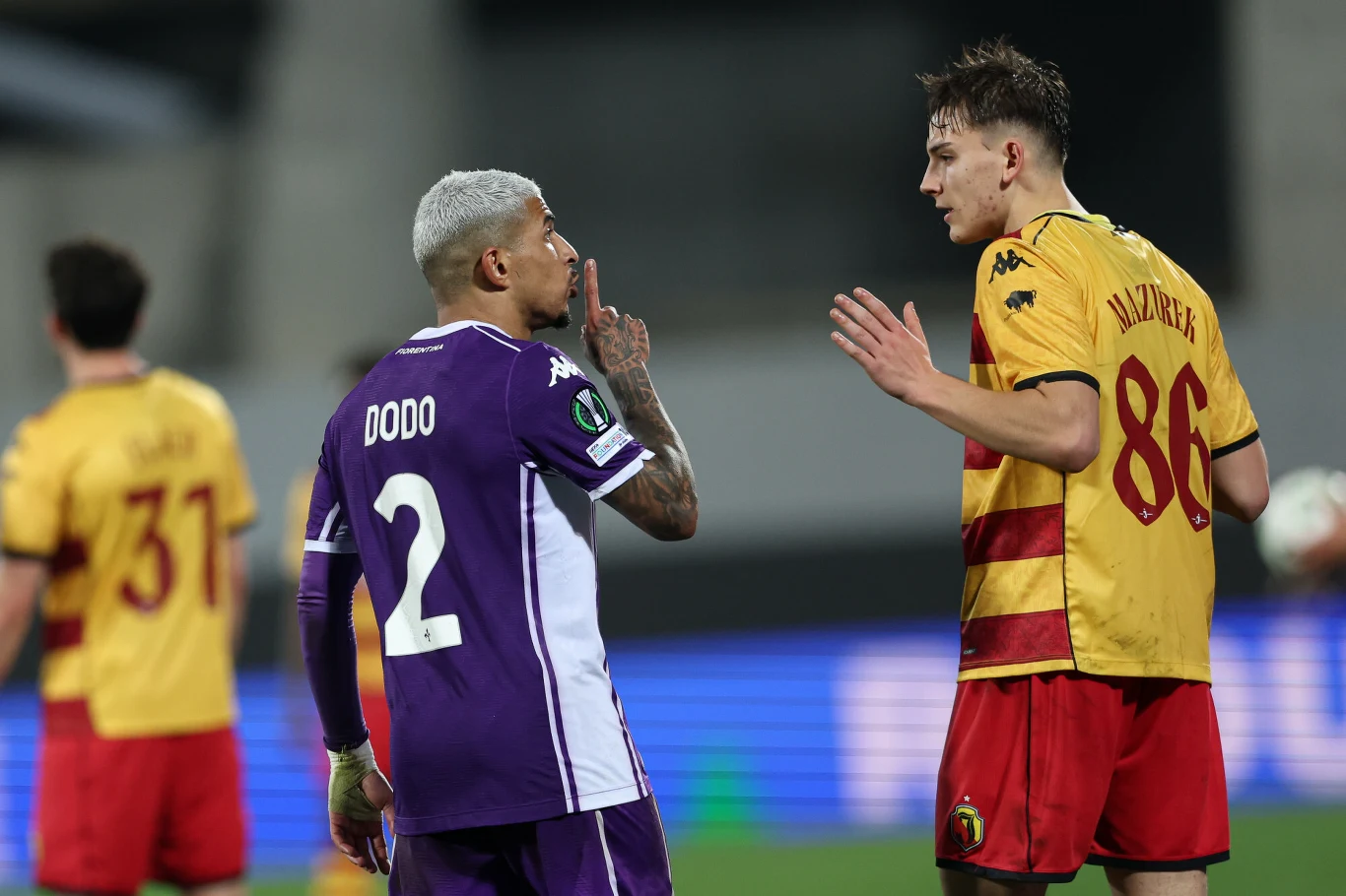 Dodo i Bartosz Mazurek podczas meczu Ligi Konferencji Fiorentina - Jagiellonia Białystok Dwóch piłkarzy stoi twarzą w twarz na boisku, jeden z nich, ubrany w fioletowy strój z numerem 2, gestykuluje w stronę przeciwnika w żółto-czerwonym stroju z numerem 86. W tle widać innych zawodników.