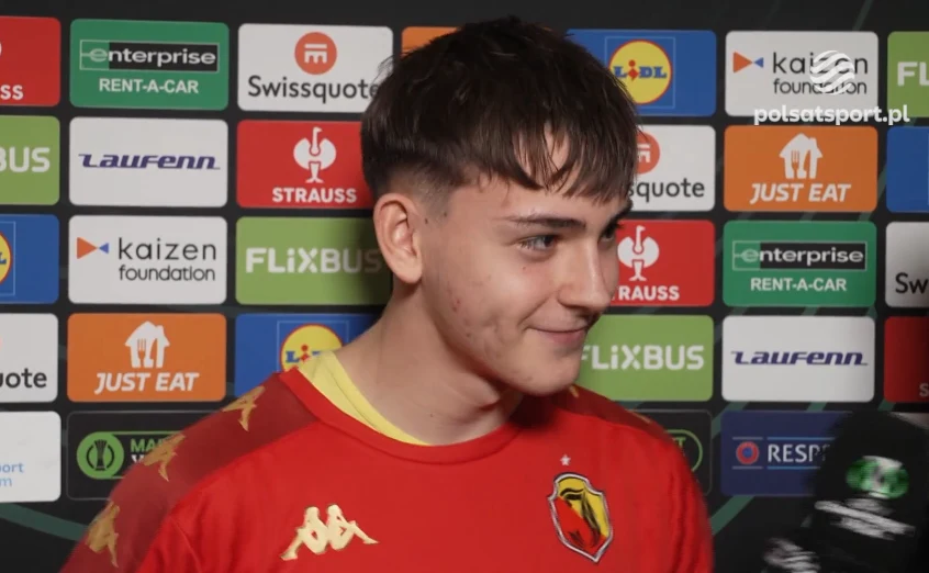 Młody mężczyzna w czerwonej sportowej koszulce z godłem drużyny udziela wywiadu, w tle tablica sponsora z wieloma logotypami różnych firm.