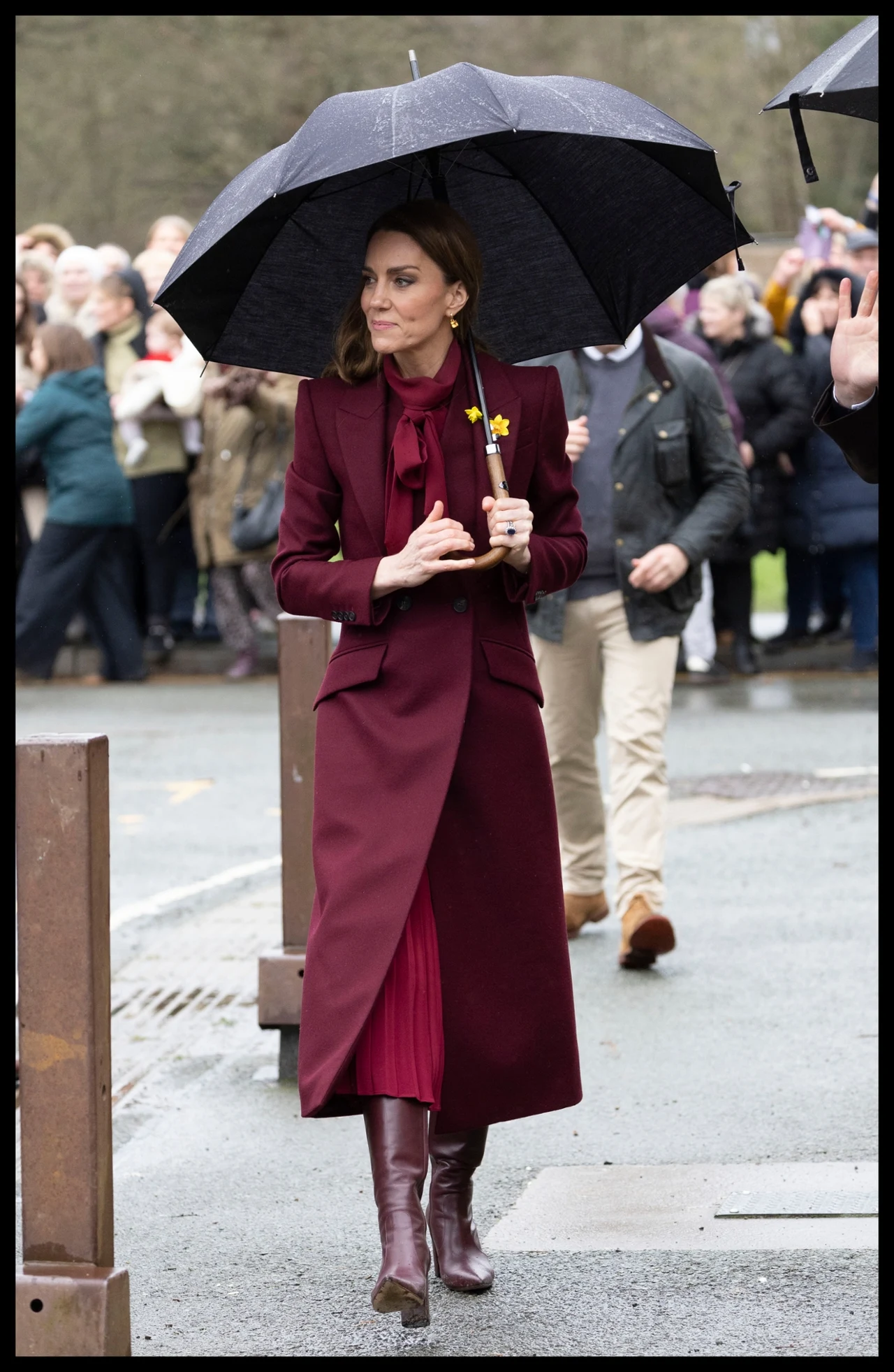 Elegancka kobieta w bordowym płaszczu i długich kozakach idzie po chodniku trzymając czarny parasol. W tle widać grupę obserwujących ludzi. Kobieta ma przypięty żółty kwiatek do płaszcza.