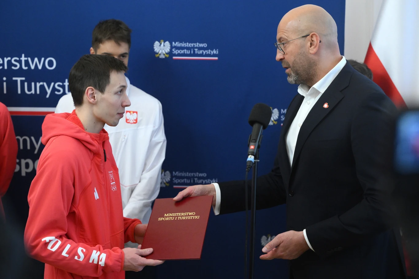 Młody mężczyzna w czerwonej bluzie z napisem POLSKA odbiera dokument od elegancko ubranego mężczyzny podczas oficjalnej ceremonii w Ministerstwie Sportu i Turystyki. W tle widoczni są inni uczestnicy wydarzenia, a także logo ministerstwa.
