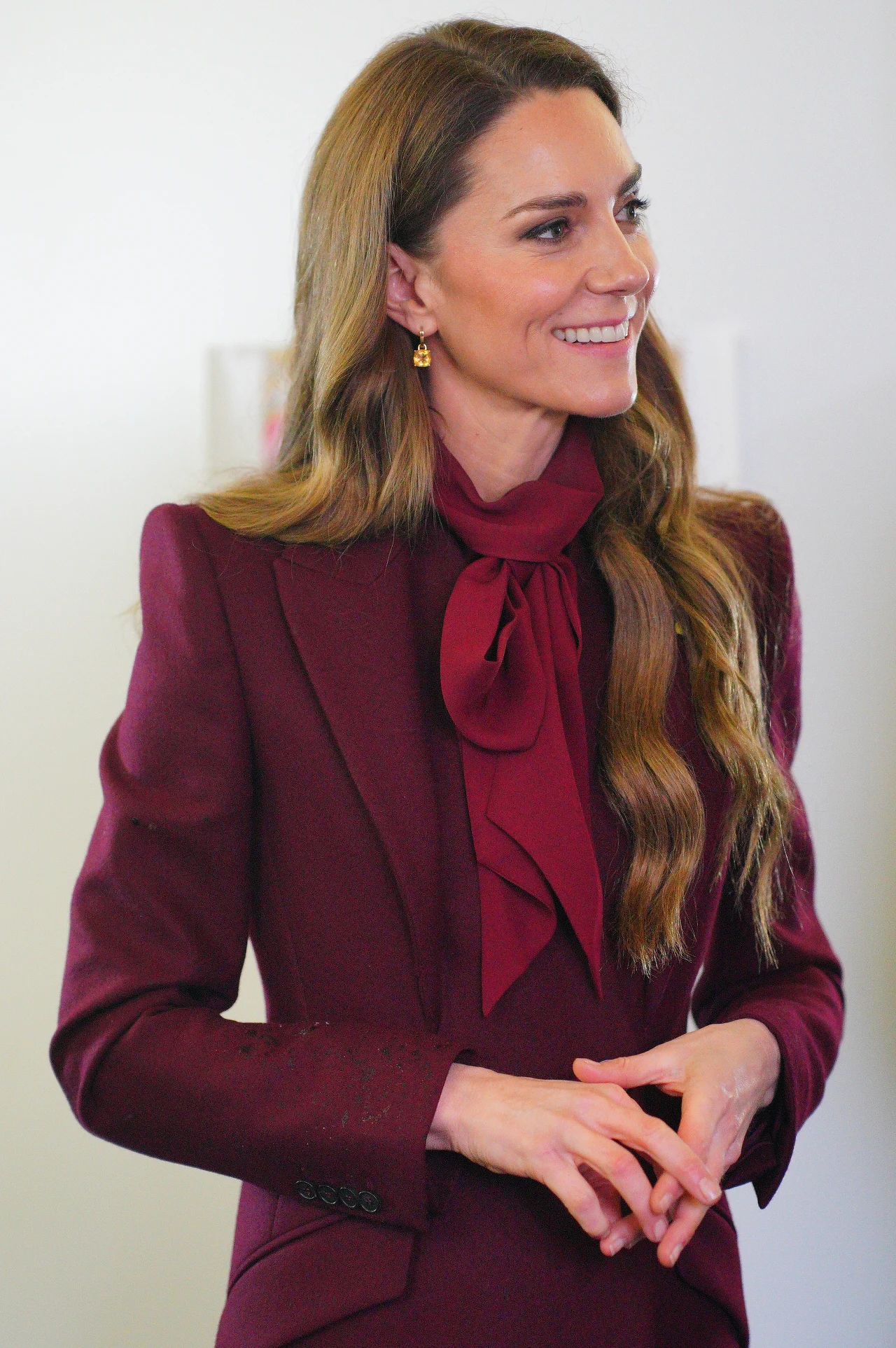 Kobieta w bordowym garniturze ze stylową apaszką, stojąca w jasnym pomieszczeniu, delikatnie uśmiechnięta, z długimi falującymi włosami i subtelną biżuterią.