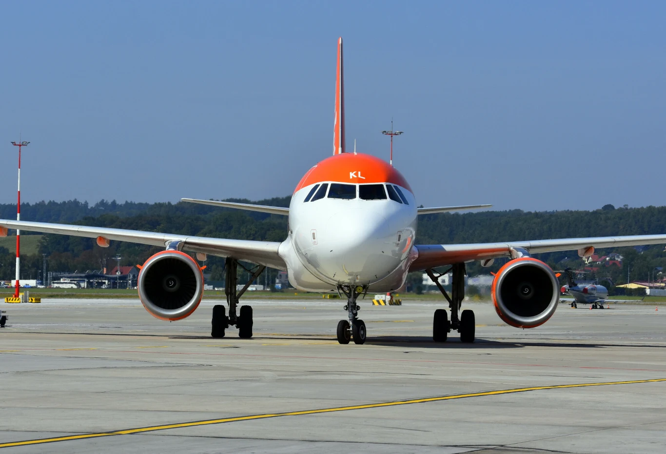 Samolot pasażerski w pomarańczowo-białych barwach stoi na płycie lotniska, widoczny jest od frontu na tle pasa startowego i niskiej zabudowy oraz drzew w oddali. Jasne niebo i przygotowany do lotu statek powietrzny dominują w kadrze.