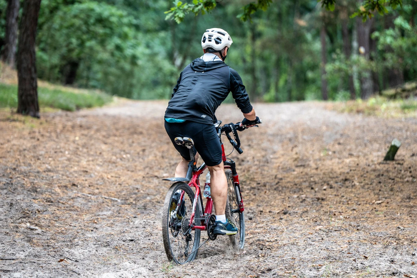 Mężczyzna w kasku rowerowym jedzie czerwonym rowerem po leśnej ścieżce otoczonej drzewami, ubrany w sportowy strój, teren piaszczysty i częściowo porośnięty trawą.