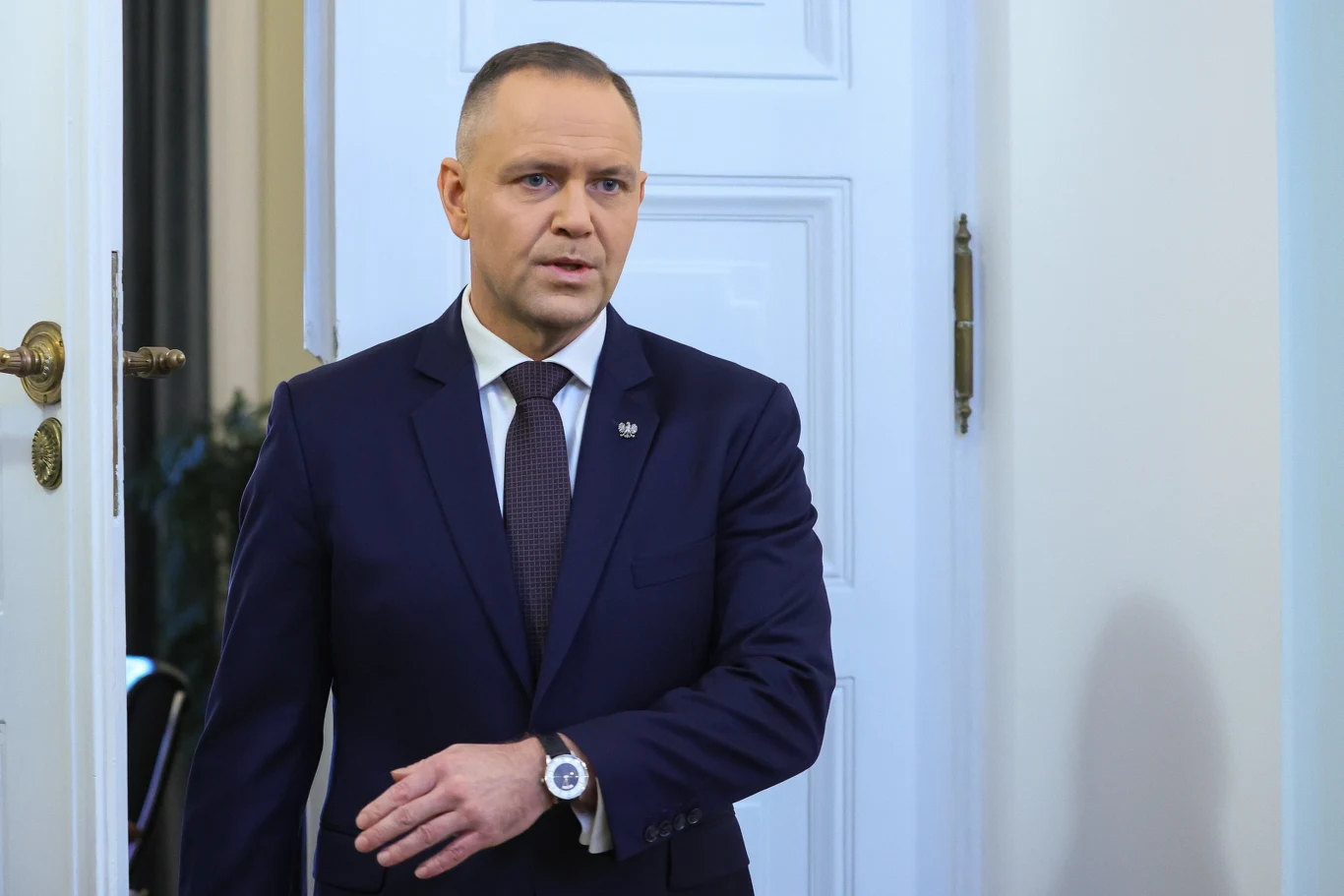 Mężczyzna w garniturze i krawacie, z przypiętym znacznikiem na klapie, stoi przy białych drzwiach, patrząc zdecydowanie przed siebie, w geście sprawdza zegarek na nadgarstku.