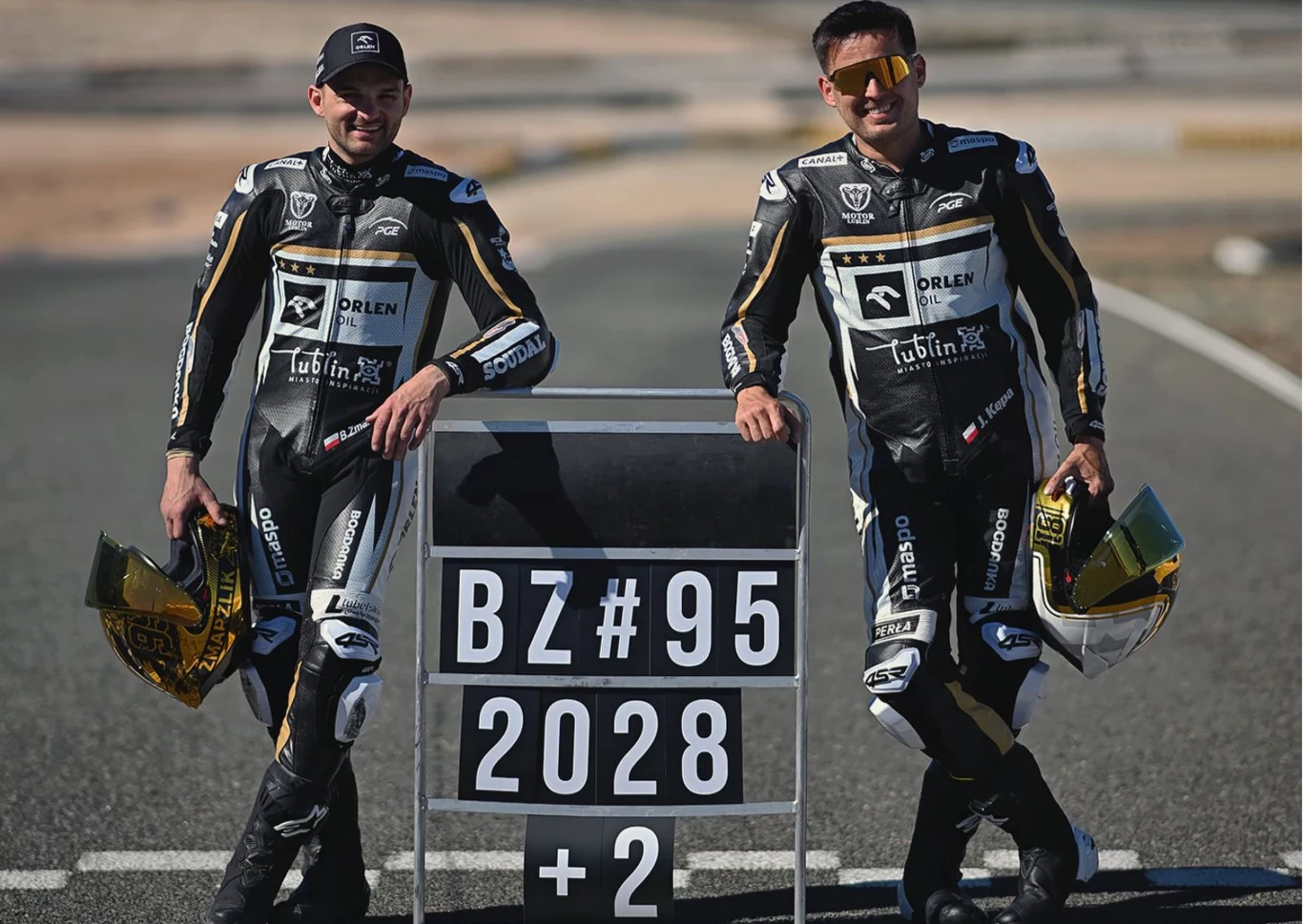 Prezes Motoru Lublin trzy lata pracował na nowy kontrakt Bartosza Zmarzlika. Dwóch motocyklistów w kombinezonach wyścigowych opiera się o tablicę wyników z numerem BZ#95, datą 2028 oraz cyfrą +2. Motocykliści trzymają kaski, stoją na torze wyścigowym.