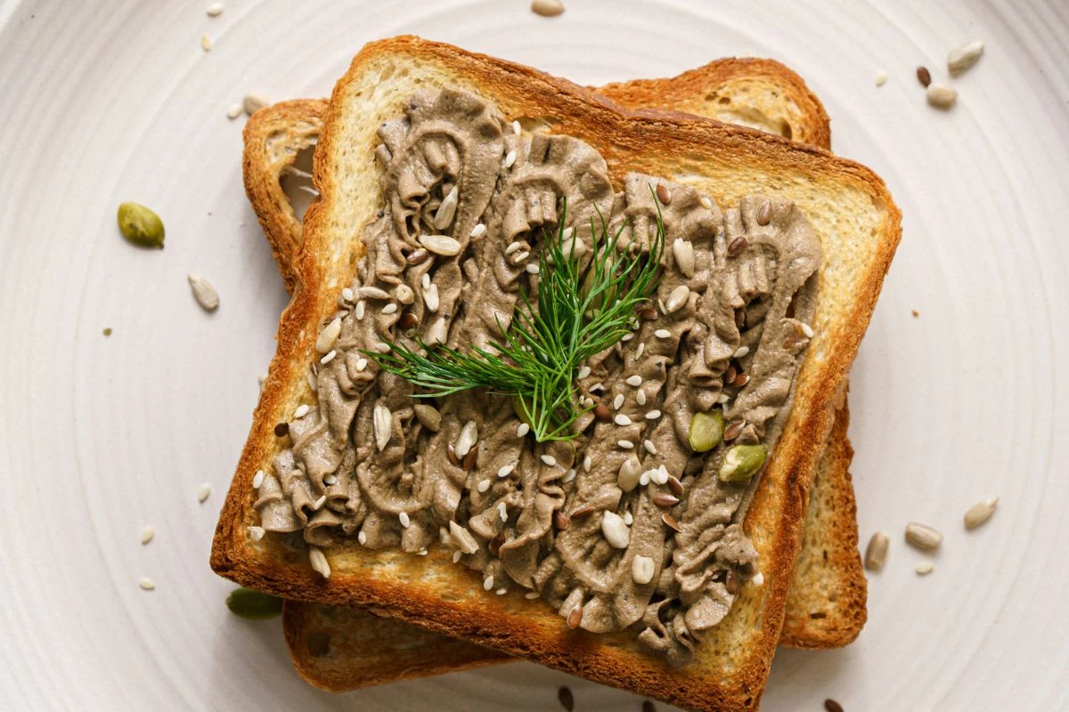 Grzanka posmarowana pasztetem, posypana ziarnami słonecznika i dyni, udekorowana gałązką koperku, umieszczona na jasnym talerzu.