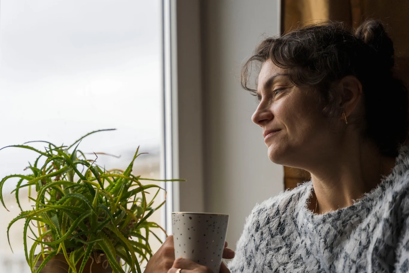Kobieta w szarym swetrze trzyma kubek i patrzy przez okno, obok na parapecie stoi zielona roślina doniczkowa.
