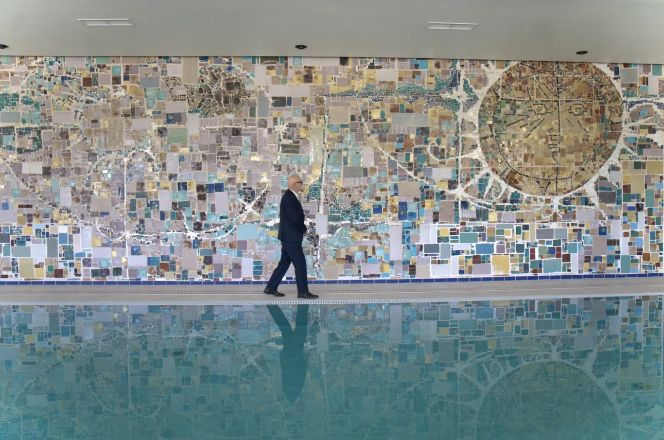Mężczyzna idzie wzdłuż dekoracyjnej ściany pokrytej kolorową mozaiką przedstawiającą abstrakcyjne wzory i dużą okrągłą mapę, poniżej widoczne jest odbicie ściany i postaci w powierzchni wody.