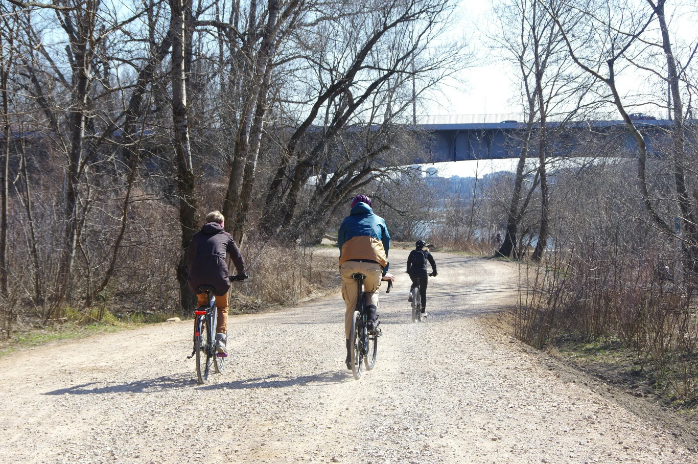 Trzy osoby jadą na rowerach szeroką, żwirową drogą przez zalesioną okolicę, widoczny wiadukt nad rzeką w tle, drzewa są jeszcze bez liści, a pogoda jest słoneczna.
