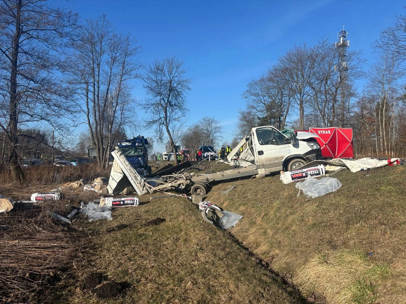 Tragiczny poranek w Małopolsce. Zdarzyły się dwa wypadki, nie żyje jedna osoba Rozbity samochód dostawczy leżący na poboczu, liczne porozrzucane towary budowlane, służby ratunkowe oraz parawan osłaniający znajdują się na miejscu zdarzenia, w tle drzewa bez liści.