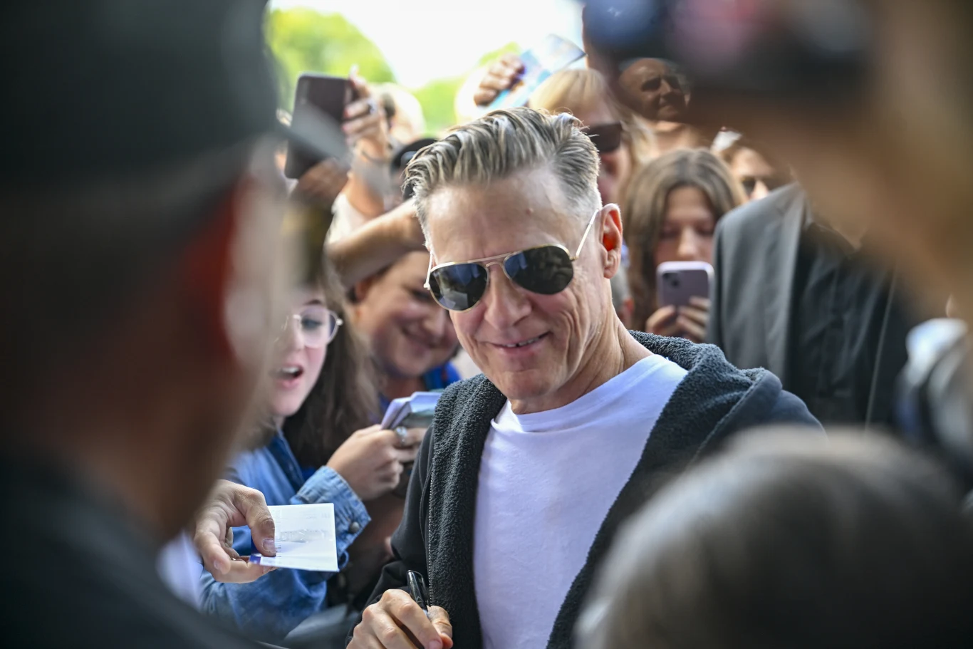 Bryan Adams regularnie odwiedza Polskę Mężczyzna w okularach przeciwsłonecznych uśmiecha się, podpisując autografy dla licznej grupy fanów, którzy trzymają telefony i kartki, próbując zbliżyć się do niego.
