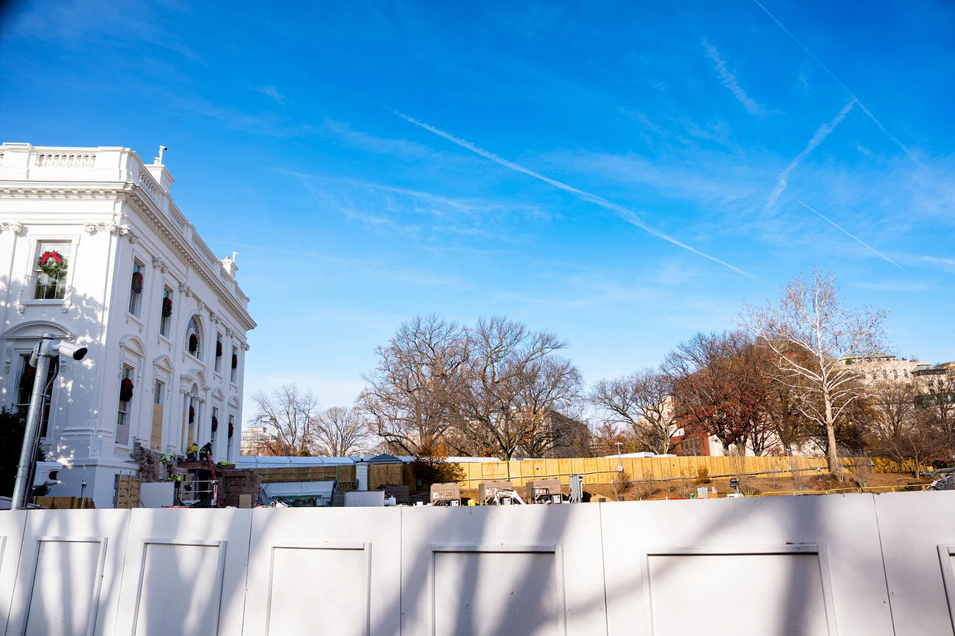 Sąd w USA orzekł, że projekt Trumpa dotyczący sali balowej może być na razie kontynuowany Biała fasada budynku w stylu klasycystycznym po lewej stronie, plac z drewnianym płotem i materiałami budowlanymi pośrodku, drzewa bez liści oraz czyste, niebieskie niebo z widocznymi smugami samolotów.