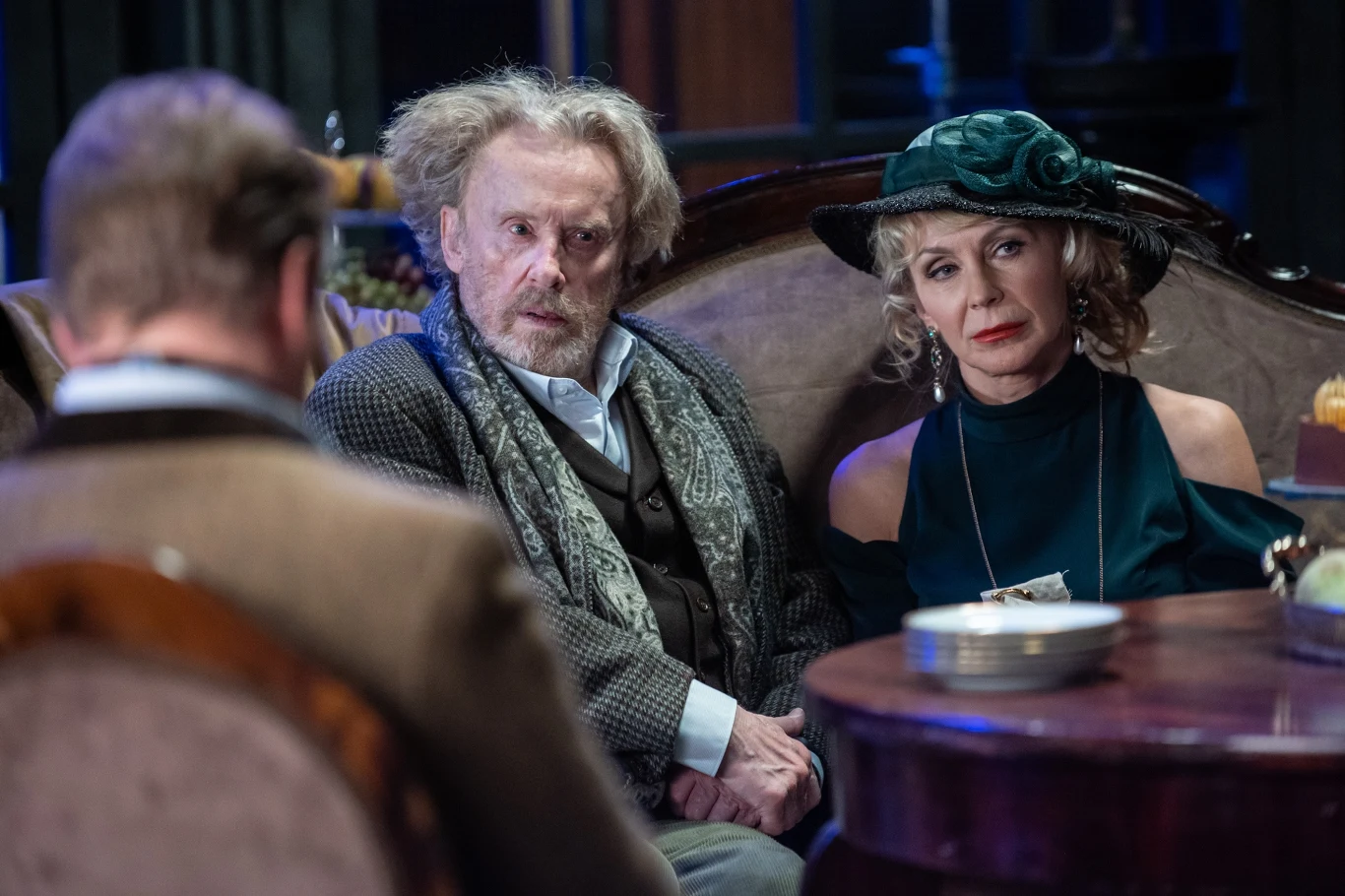 Starszy mężczyzna z nieładem we włosach i poważnym wyrazem twarzy oraz elegancko ubrana kobieta w kapeluszu siedzą przy okrągłym stole, naprzeciw mężczyzny z tyłu, w pomieszczeniu o teatralnym klimacie.