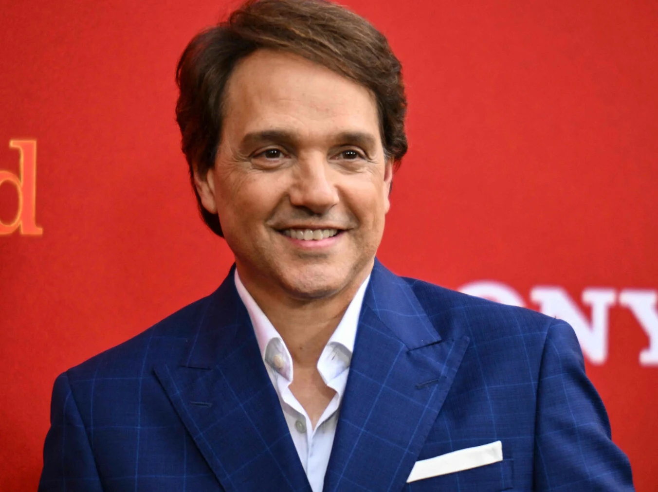 Mężczyzna w garniturze w kratę ze śnieżnobiałą koszulą, uśmiechnięty, stoi na czerwonym tle z widocznymi fragmentami białych napisów.