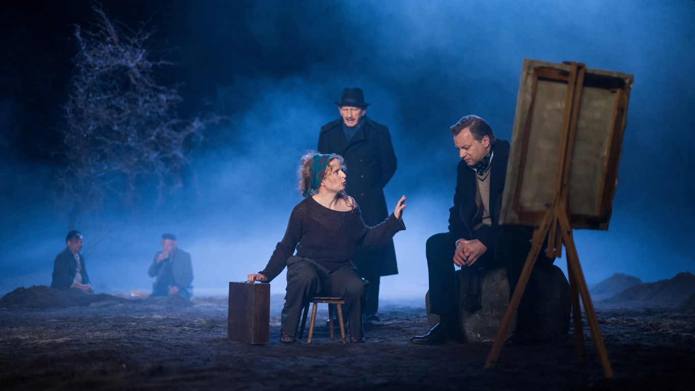 Scena teatralna w plenerze, artystka siedzi przed sztalugą i rozmawia z mężczyzną, w tle dwaj mężczyźni siedzą na ziemi, całość oświetlona jest niebieskim światłem, w tle widać suche drzewo i mgłę tworzącą atmosferę tajemniczości.