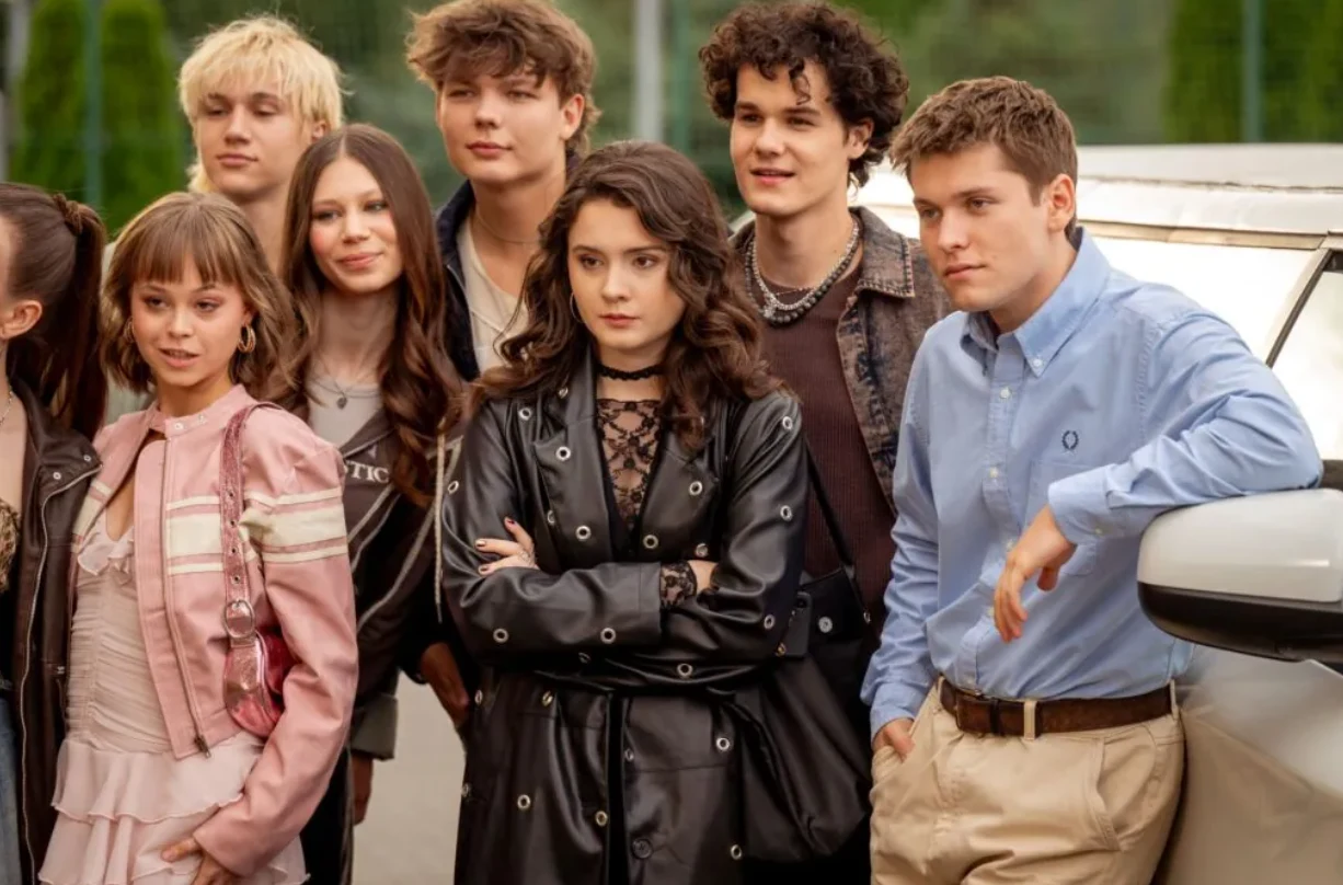 "Piep*zyć Mickiewicza 3" Grupa młodych osób stoi razem na zewnątrz, większość z nich patrzy w jednym kierunku, niektóre uśmiechnięte, jedna dziewczyna w centrum z poważnym wyrazem twarzy, nieformalna atmosfera, obok samochód.