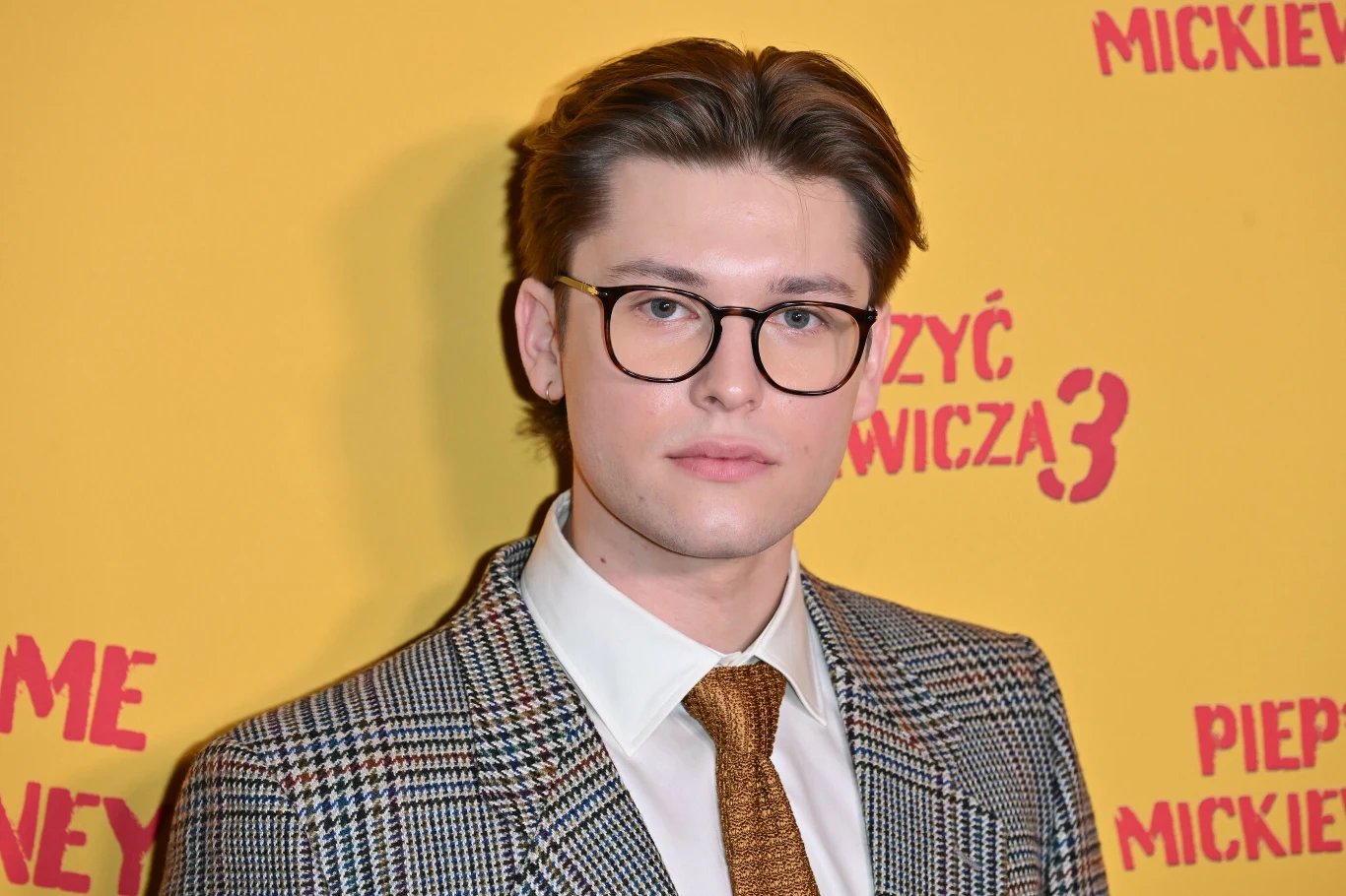 Aleksander Idzi Młody mężczyzna w okularach, ubrany w elegancki garnitur w kratę z białą koszulą i brokatowym krawatem, stoi na tle żółtej ścianki z czerwonymi napisami.