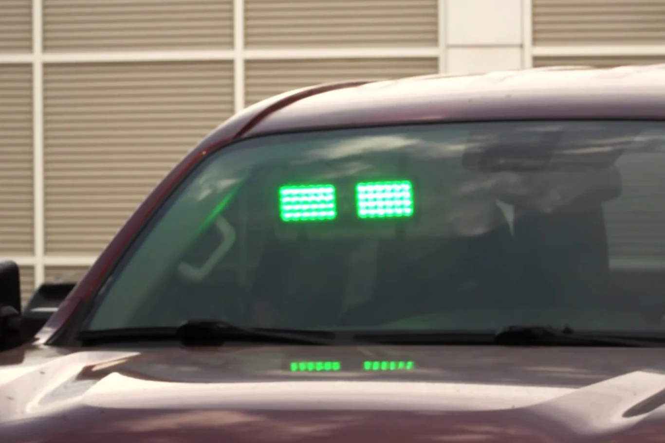 Czerwony samochód z dwoma jasnymi, zielonymi panelami świetlnymi umieszczonymi na przedniej szybie, na tle budynku z oknami osłoniętymi żaluzjami.