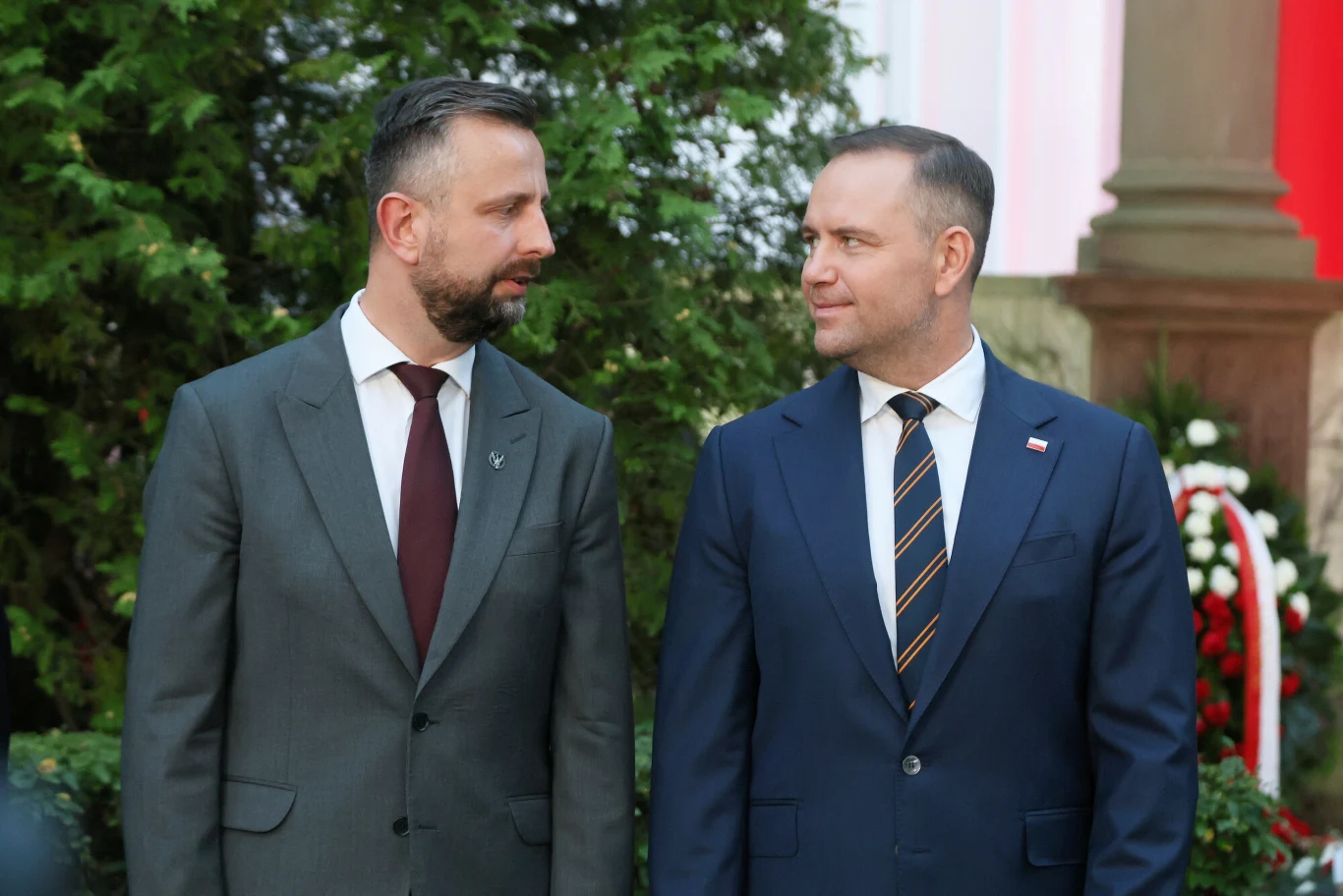 Wicepremier i szef MON Władysław Kosiniak-Kamysz i prezydent Karol Nawrocki Dwóch mężczyzn w garniturach stoi na zewnątrz na tle zielonych krzewów oraz biało-czerwonej dekoracji, rozmawiając ze sobą, obaj patrzą sobie w oczy.