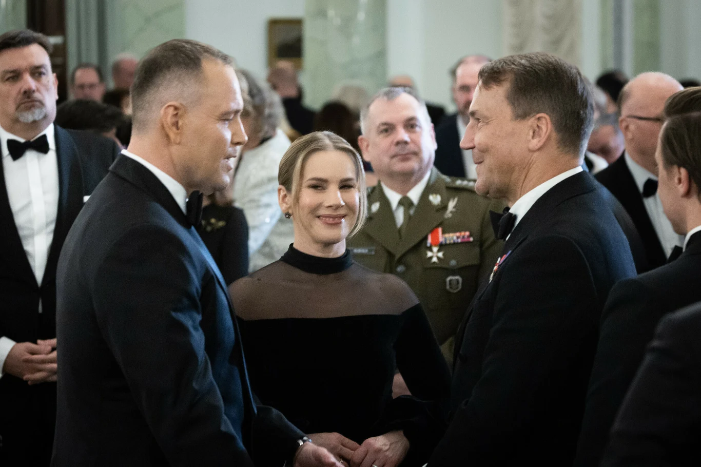 Grupa elegancko ubranych osób podczas oficjalnego spotkania, dwaj mężczyźni w garniturach rozmawiają ze sobą, obok nich kobieta w czarnej sukni, w tle mężczyzna w mundurze wojskowym oraz inni uczestnicy przyjęcia.