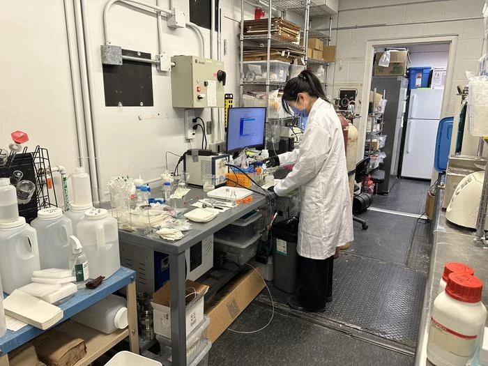 Osoba w białym fartuchu laboratoryjnym wykonuje eksperymenty lub badania przy stanowisku wyposażonym w sprzęt laboratoryjny, komputery, butelki oraz naczynia z odczynnikami chemicznymi, a otoczenie sugeruje nowoczesne laboratorium badawcze.