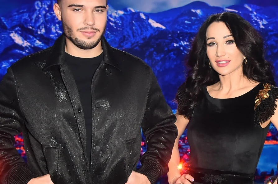 Leon Myszkowski z Justyną Steczkowską Para elegancko ubranych osób pozuje na tle górskiego krajobrazu, kobieta w czarnej sukni z ozdobnym ramieniem, mężczyzna w ciemnej kurtce, oboje uśmiechnięci i swobodnie stojący obok siebie.