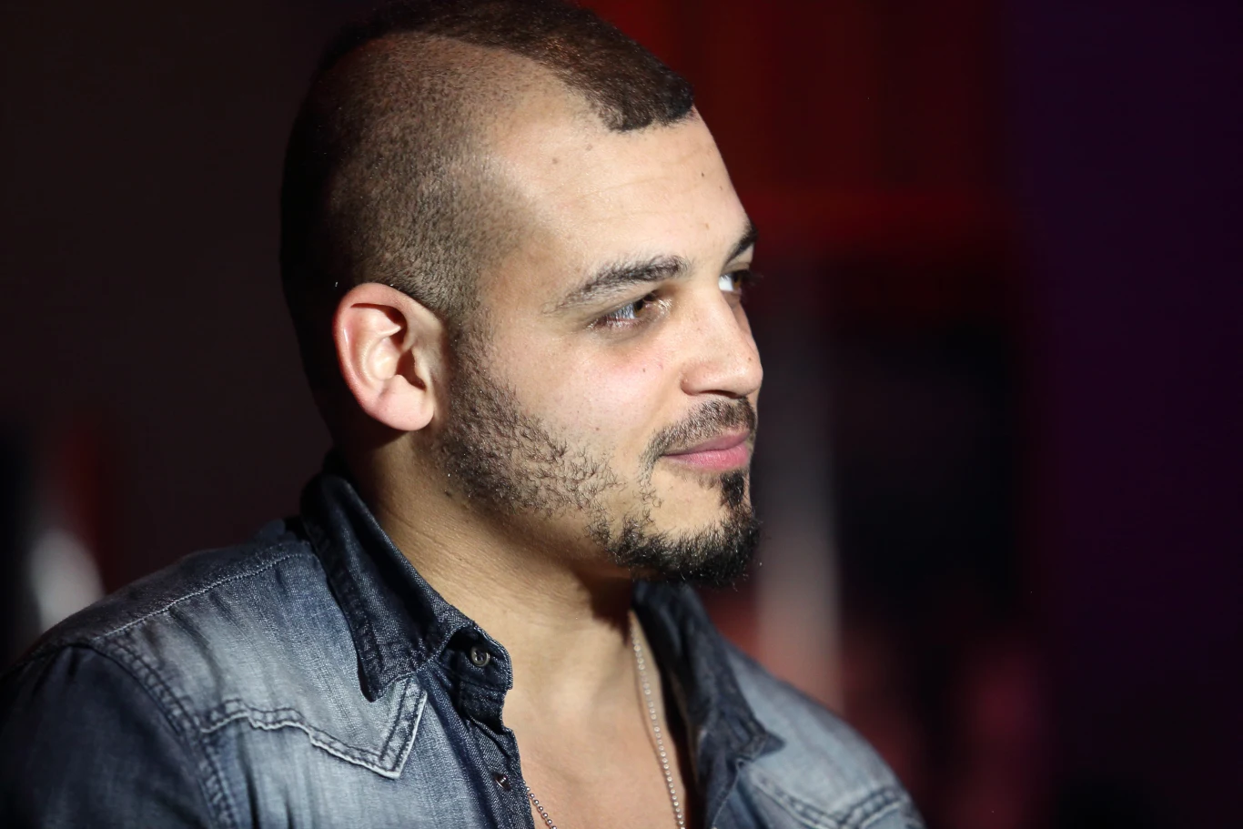 Damian Ukeje Mężczyzna z ogoloną głową i delikatnym zarostem ubrany w koszulę jeansową, patrzący w bok, o łagodnym wyrazie twarzy, sfotografowany na neutralnym tle.