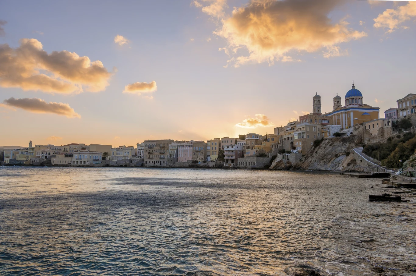 Widok nadmorskiego miasta w blasku zachodzącego słońca, po prawej stronie wznoszą się historyczne budynki i duża kopuła kościoła z dwiema wieżami, w tle rozproszone chmury podświetlone przez promienie słońca.