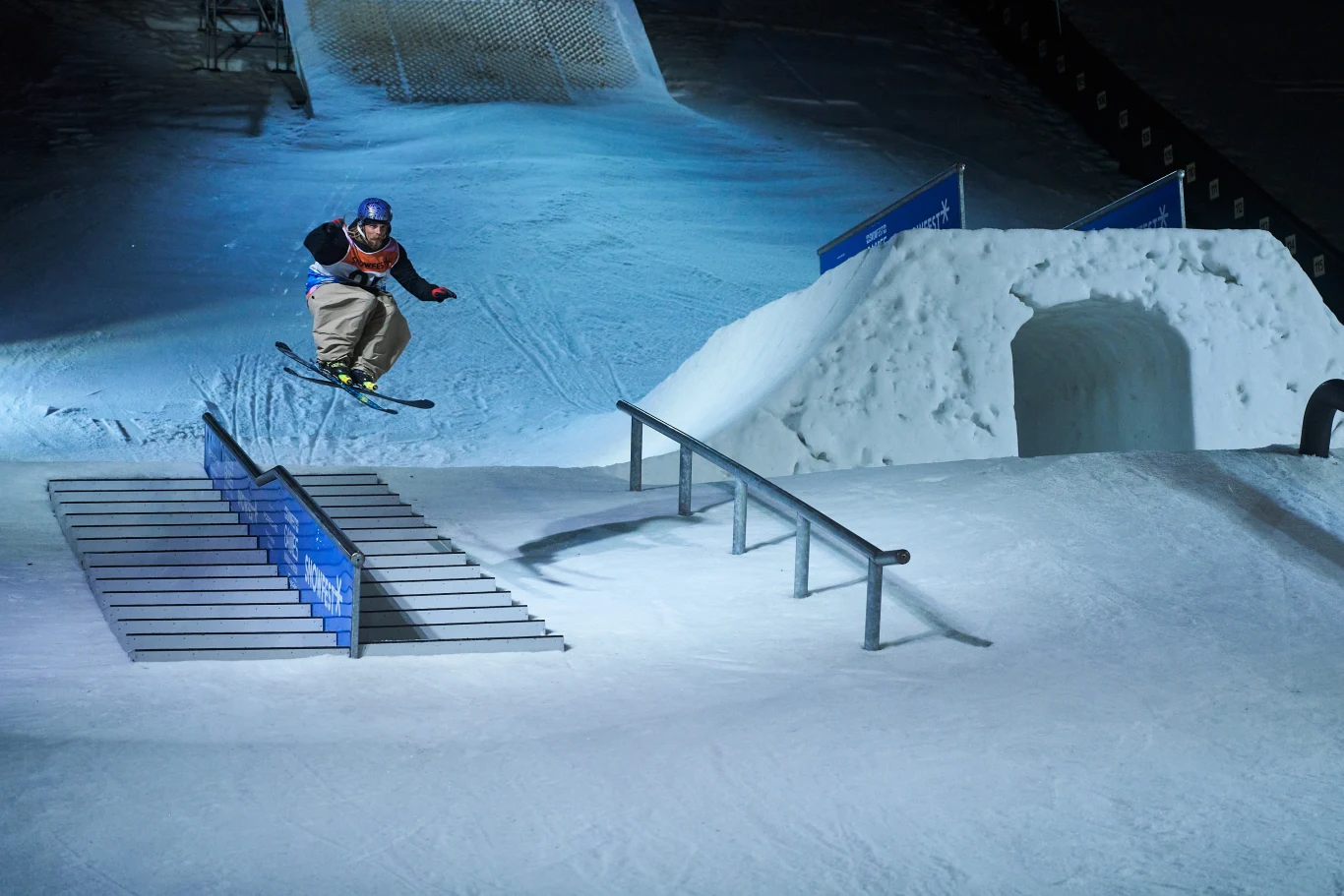 Narciarz wykonujący skok oraz trik na specjalnie przygotowanym torze snowparku podczas nocnej jazdy, otoczony śniegiem i konstrukcjami służącymi do wykonywania ewolucji.