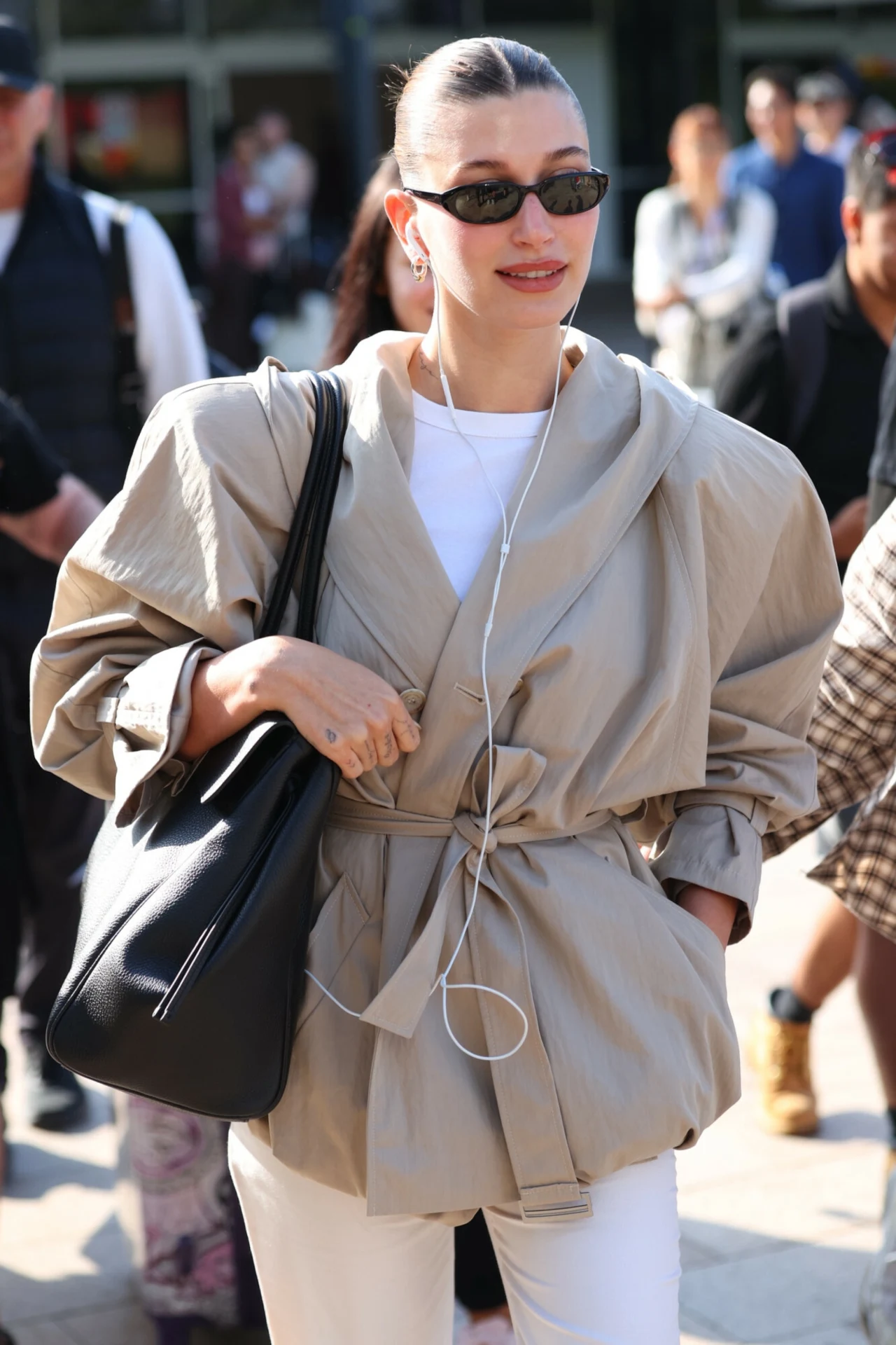 Kobieta związała włosy w kok, nosi stylowe okulary przeciwsłoneczne, beżowy oversize’owy płaszcz z paskiem, białe spodnie oraz duży czarny torebkę na ramieniu, słucha muzyki przez słuchawki, stoi w tłumie osób na ulicy.