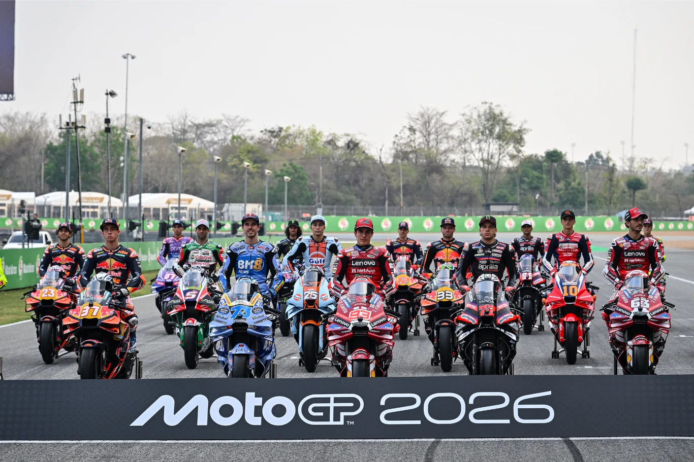 Zawodnicy MotoGP startujący w sezonie 2026. Czołowi zawodnicy MotoGP ustawieni ze swoimi motocyklami na torze wyścigowym, wszyscy ubrani w profesjonalne kombinezony wyścigowe, na pierwszym planie duża tablica z napisem MotoGP 2026
