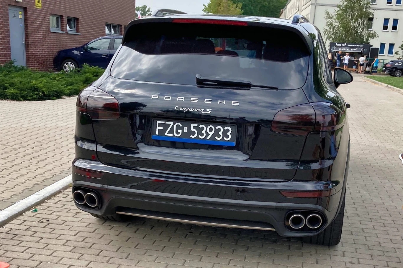 Czarny samochód Porsche Cayenne S zaparkowany na kostce brukowej, widoczny od tyłu, z wyraźnym numerem rejestracyjnym i czterema końcówkami układu wydechowego, w tle budynki oraz zaparkowane pojazdy.