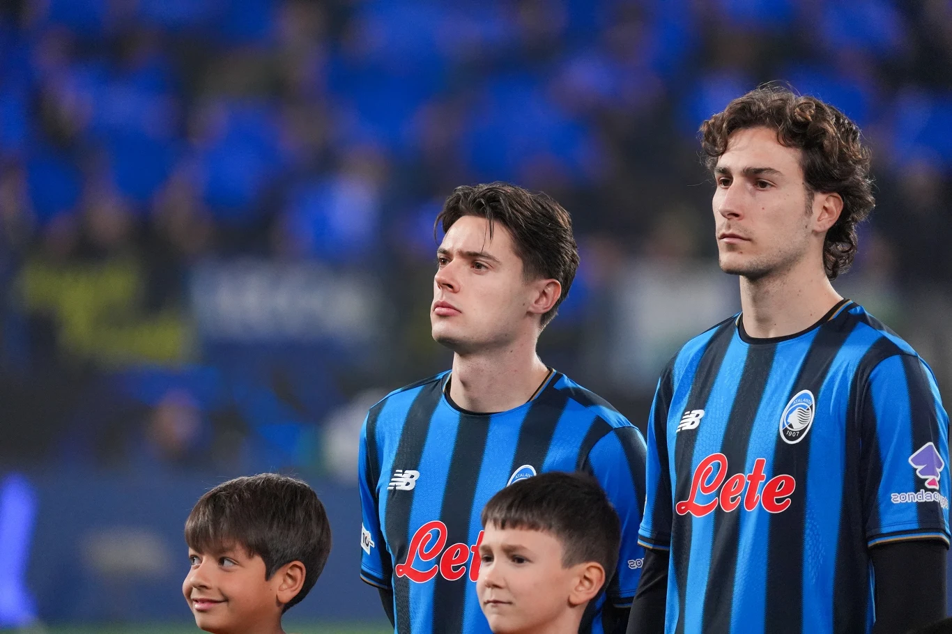 Nicola Zalewski w z lewej Dwóch piłkarzy w pasiastych, niebiesko-czarnych koszulkach drużyny stoi obok siebie, z przodu znajdują się dwaj młodzi chłopcy, tło rozmyte, pełne niebieskich barw trybun.