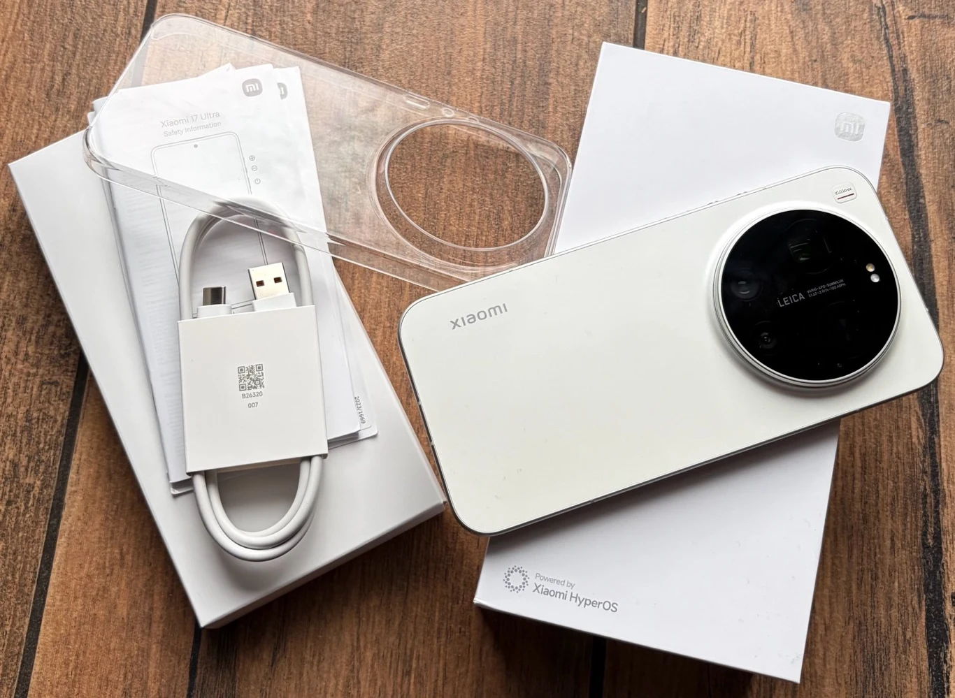 Biały smartfon Xiaomi z dużym okrągłym aparatem, przezroczyste etui, kabel USB z adapterem oraz oryginalne opakowania produktu ułożone na drewnianej powierzchni.