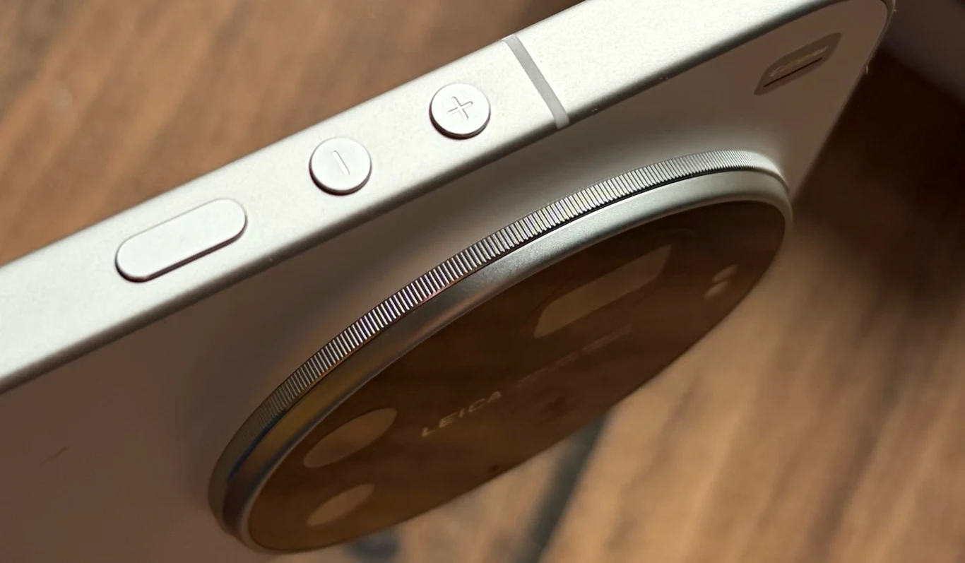 Fragment smartfona z wyraźnie widocznym modułem aparatu fotograficznego oraz przyciskami regulacji głośności, osadzony na tle drewnianej powierzchni.